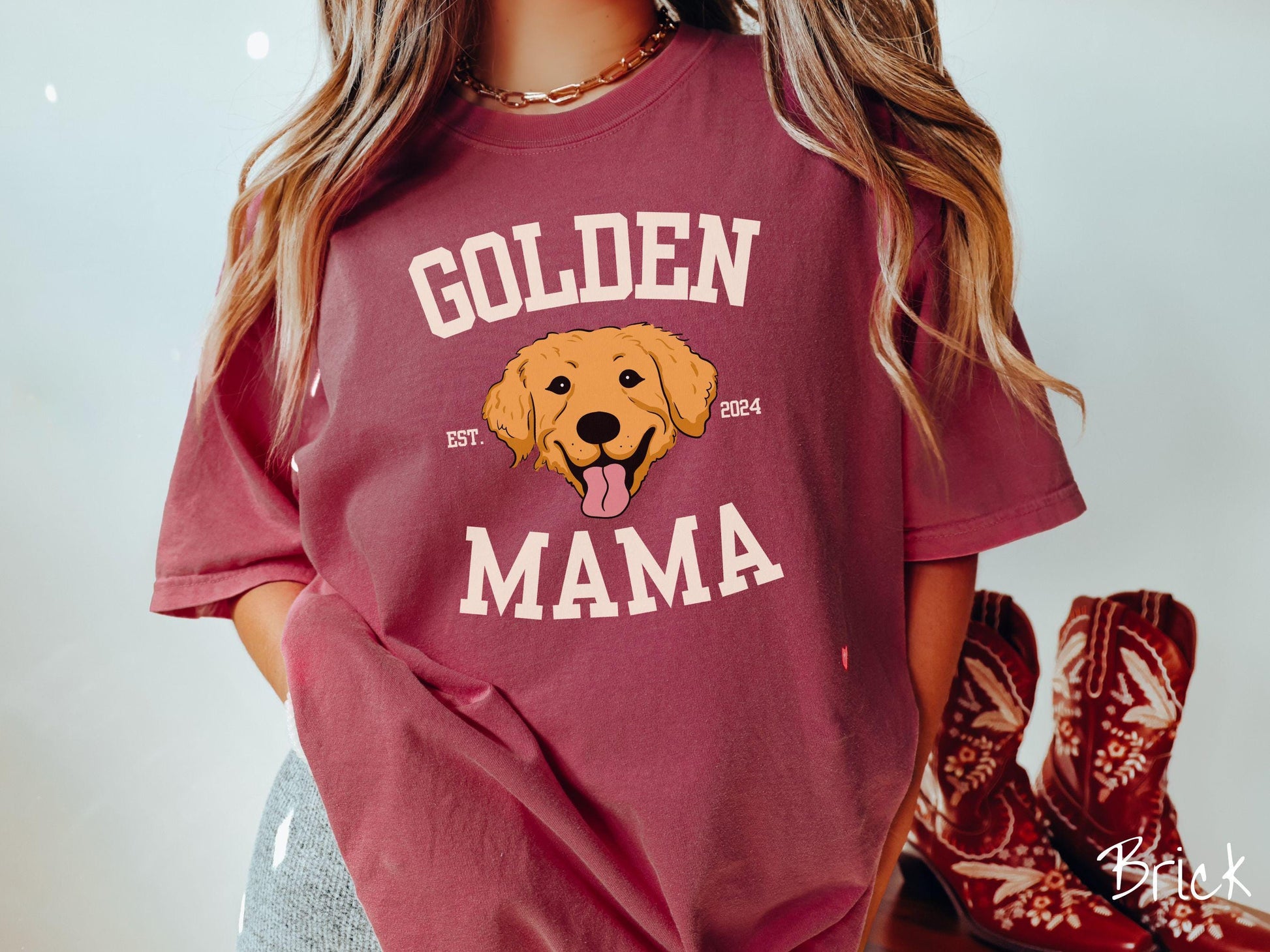 A vintage Comfort Colors t-shirt features a design with the words Golden Mama in a classic university-style font, framing an adorable black and orange Golden Retriever with its tongue out. The playful touch of EST. 2024 adds charm to the design.
