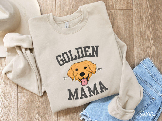 A vintage Gildan sweatshirt features a design with the words Golden Mama in a classic university-style font, framing an adorable black and orange Golden Retriever with its tongue out. The playful touch of EST. 2024 adds charm to the design.