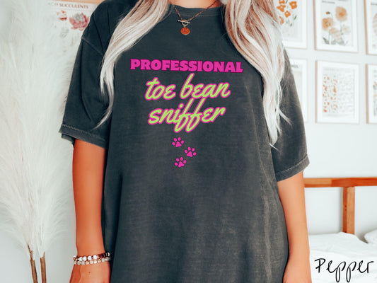 A vintage Comfort Colors t-shirt perfect for dog lovers who can’t resist those adorable toe beans! It features the funny text Professional Toe Bean Sniffer in bold pink and green fluorescent font, and below the text are three pink dog paw prints.
