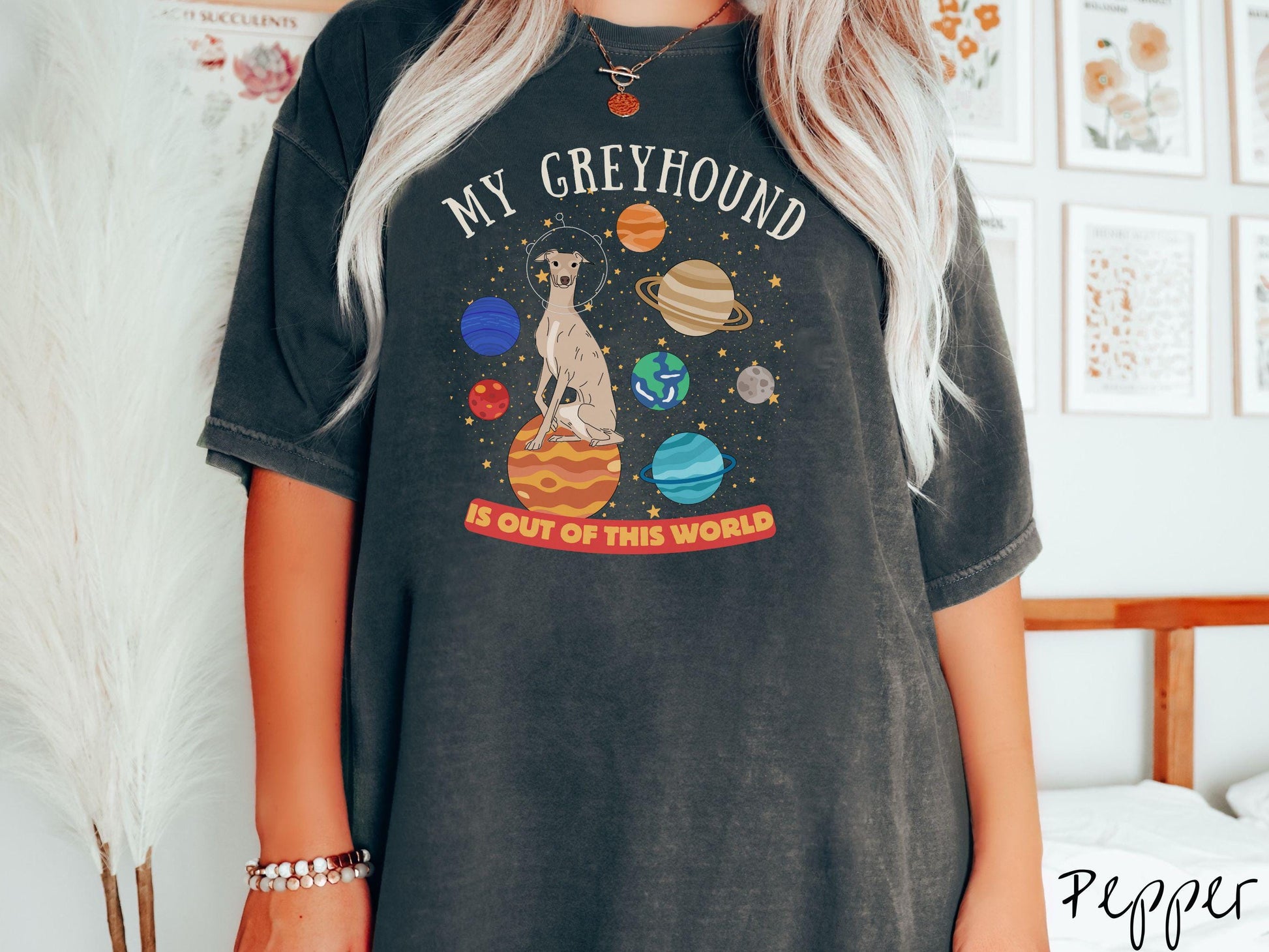 A vintage Comfort Colors t-shirt featuring the text My Greyhound is Out of This World. The design showcases a Greyhound sitting proudly atop Jupiter, surrounded by a vibrant solar system filled with colorful planets and twinkling stars.