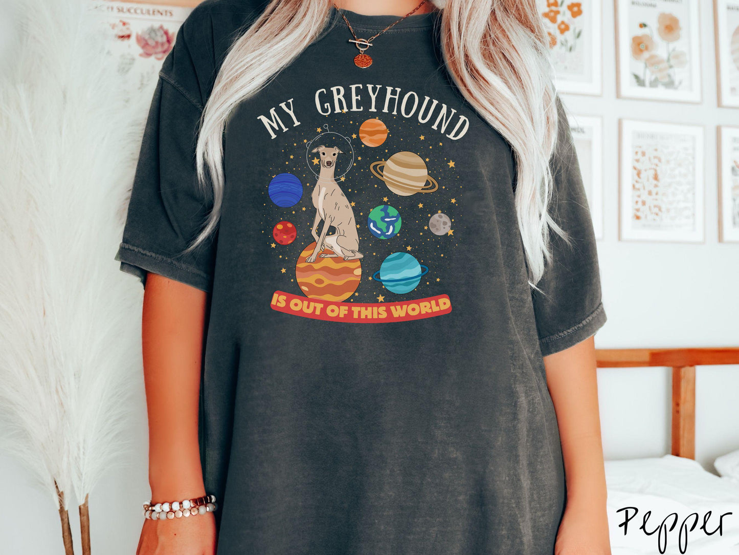 A vintage Comfort Colors t-shirt featuring the text My Greyhound is Out of This World. The design showcases a Greyhound sitting proudly atop Jupiter, surrounded by a vibrant solar system filled with colorful planets and twinkling stars.
